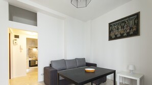 A modern living room with a dark sofa, a black table, and a white side table. A painting is on the wall, and a kitchen is visible in the background. - Studio Flat in a Quiet Place (Milan)
