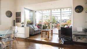 A modern living room with a glass dining table, a sofa, a TV, and a large window with a view of greenery. - The Traveller's Terrace (Milan)