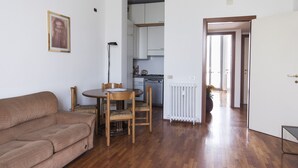 A living room with a brown sofa, wooden dining table, and chairs. There is a kitchen area with cabinets and a stove. A radiator is visible on the wall. - Bright & Cozy Flat near Navigli, Iulm, Tortona (Milan)