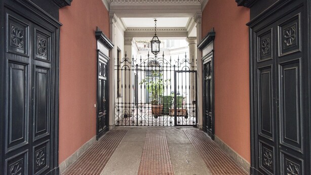 A corridor with black ornate doors and a wrought iron gate leading to a courtyard.