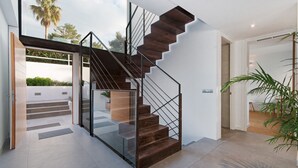 Una escalera moderna con barandillas de vidrio y peldaños de madera, que lleva a un espacio al aire libre con vistas a palmeras.