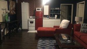 Een knus woongedeelte met een keukenhoek, inclusief een rode koelkast, een witte kookplaat en een magnetron. Er zijn twee rode banken en een glazen salontafel. De kamer heeft een donkerblauwe accentmuur en een kamerkleed met patroon op de vloer.
