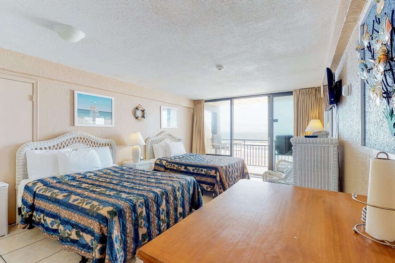 A hotel room with two beds, a wooden table, a balcony with a view, and a television on the wall.