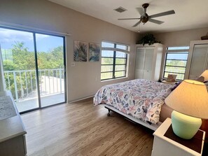 Ein Schlafzimmer mit Bett, Deckenventilator, Fenster mit Blick auf Grünflächen und einem Balkon.
