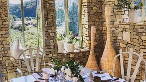Un coin repas rustique avec des murs en pierre, de grandes fenêtres et une table dressée pour le petit-déjeuner, avec divers plats et boissons.