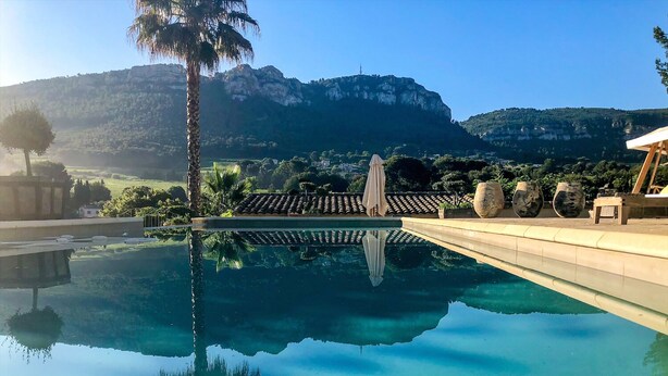 Une piscine aux eaux cristallines, avec une chaîne de montagnes en arrière-plan, un palmier et une structure surmontée d’un toit en tuiles.