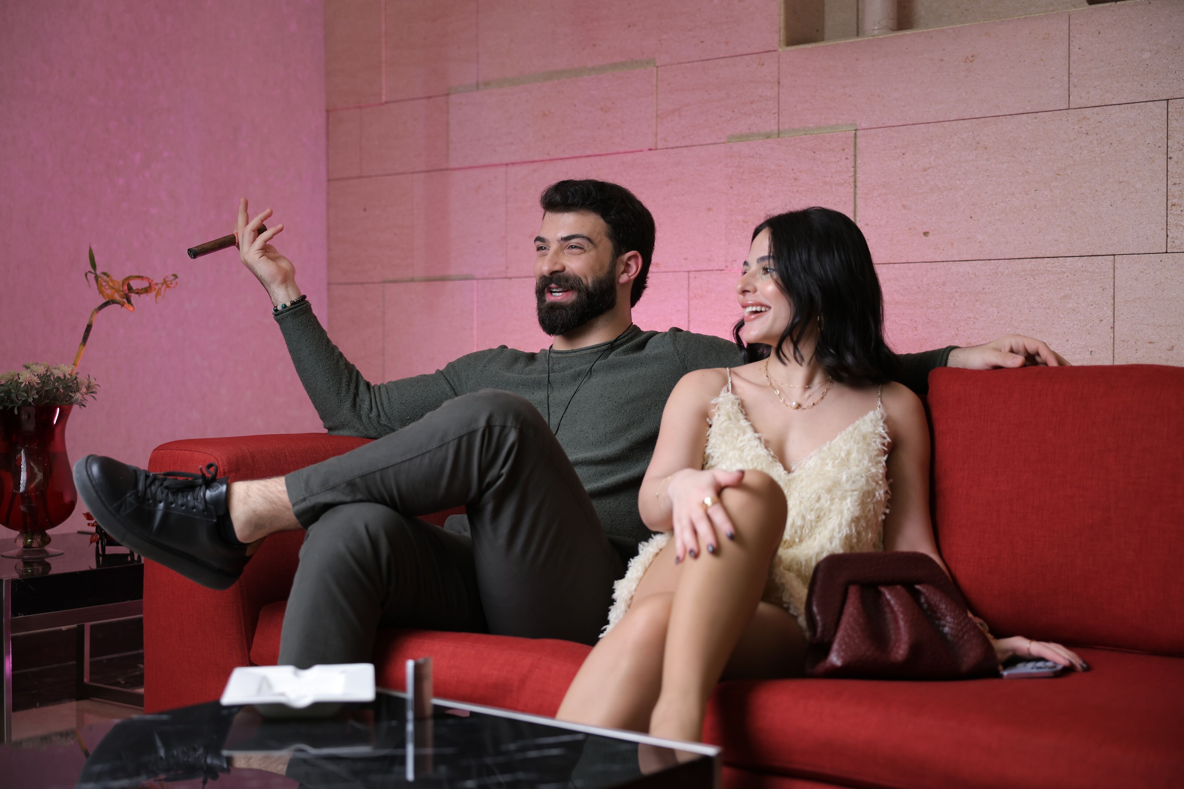 A couple sitting on a red couch in a room with a pink wall and a vase of flowers.
