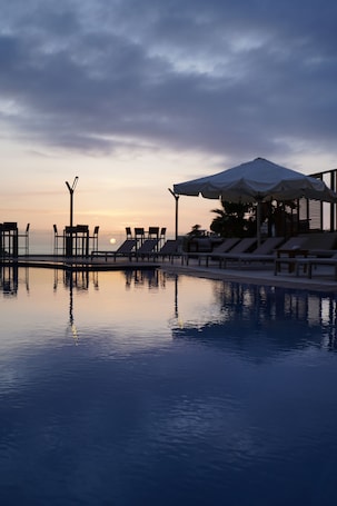 A serene pool area with lounge chairs and umbrellas, overlooking a calm body of water and a sunset.