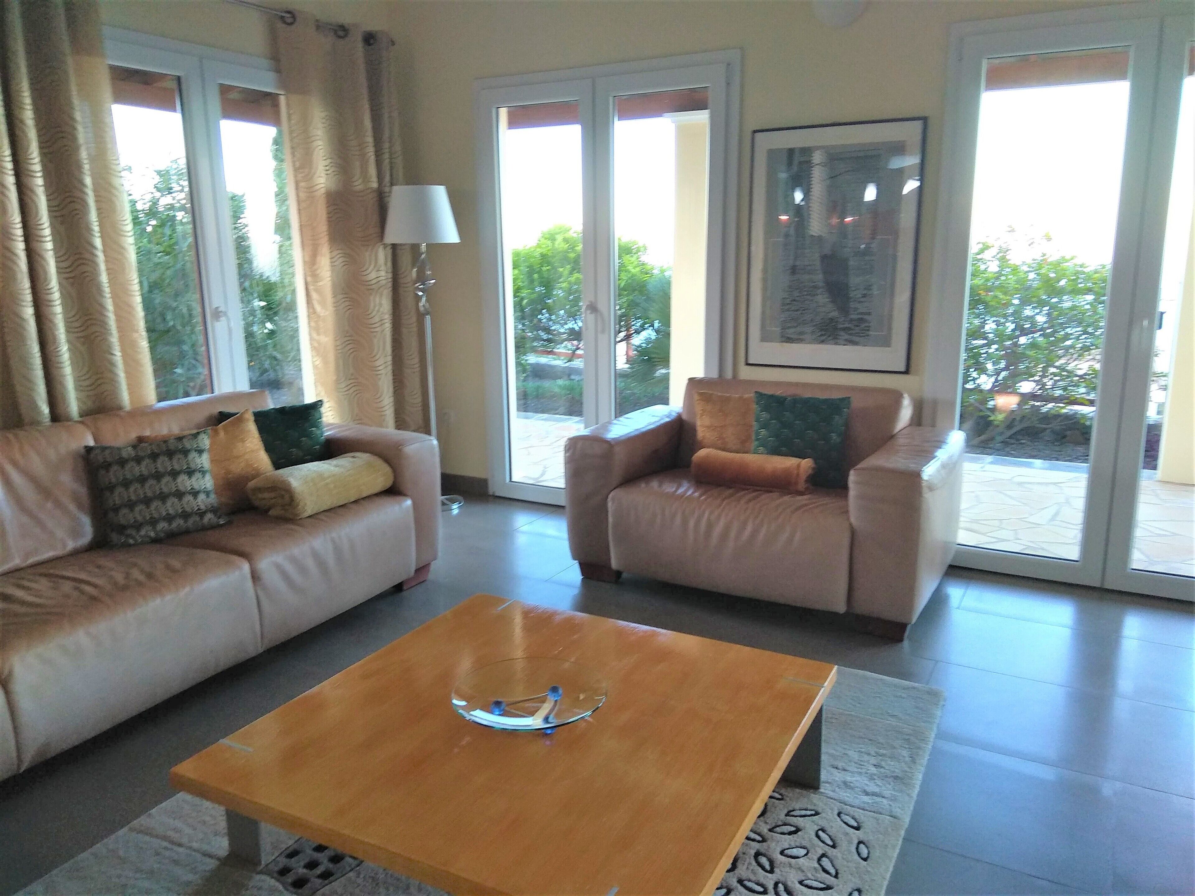 Una sala de estar con un sofá color beige, una mesa de centro de madera y una silla con cojines. Hay una lámpara de pie y un ventanal con cortinas.