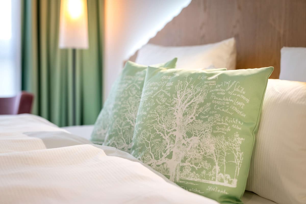 Un lit avec une literie blanche et des oreillers verts ornés de motifs d’arbres blancs.