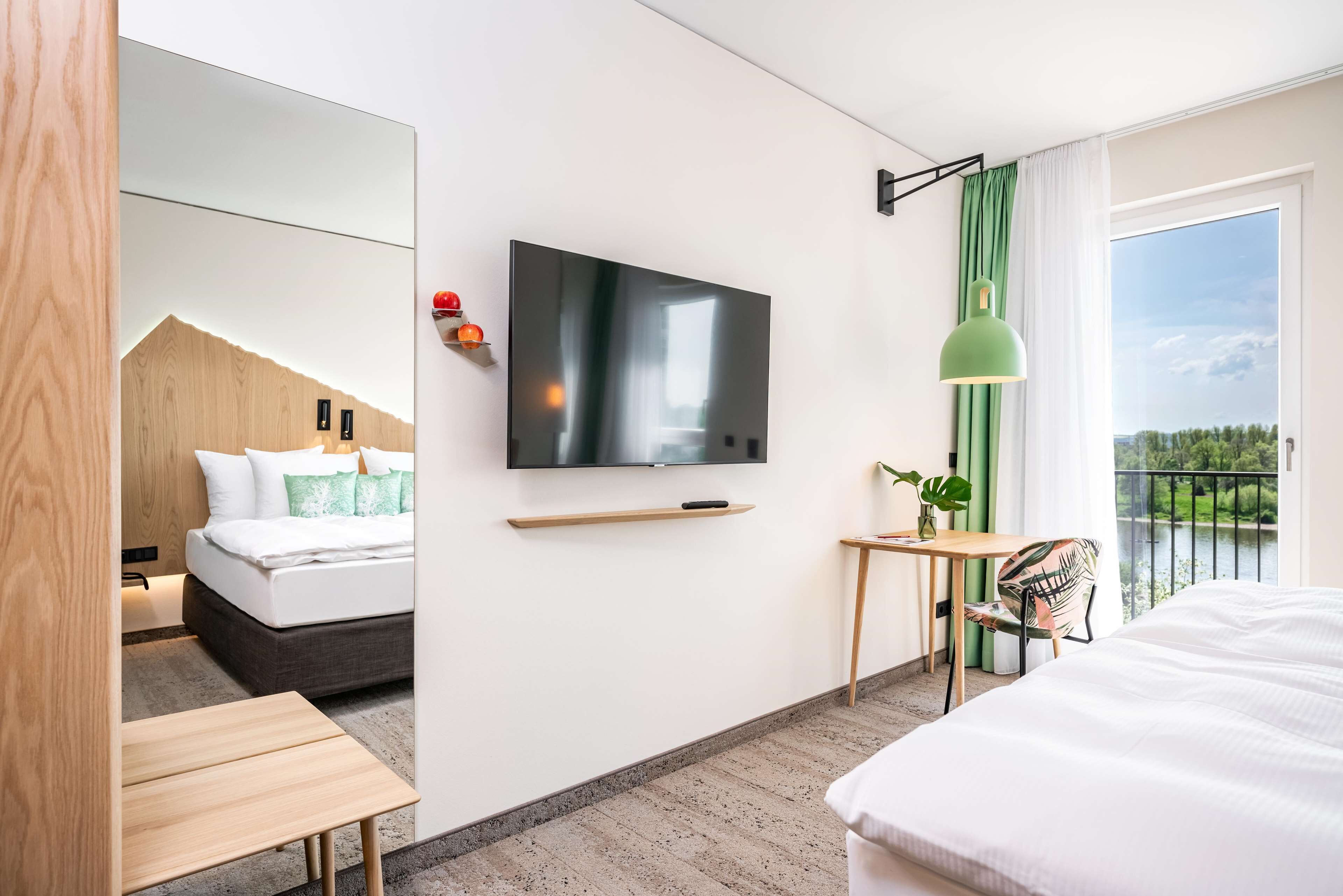 Una habitación de hotel moderna con una cama grande, un televisor montado en la pared, una mesita, una silla y vistas a un balcón con barandilla.