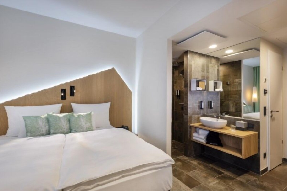 Une chambre d’hôtel moderne avec un grand lit, une tête de lit en bois, une salle de bain dotée d’un grand miroir, d’un lavabo et d’une douche.