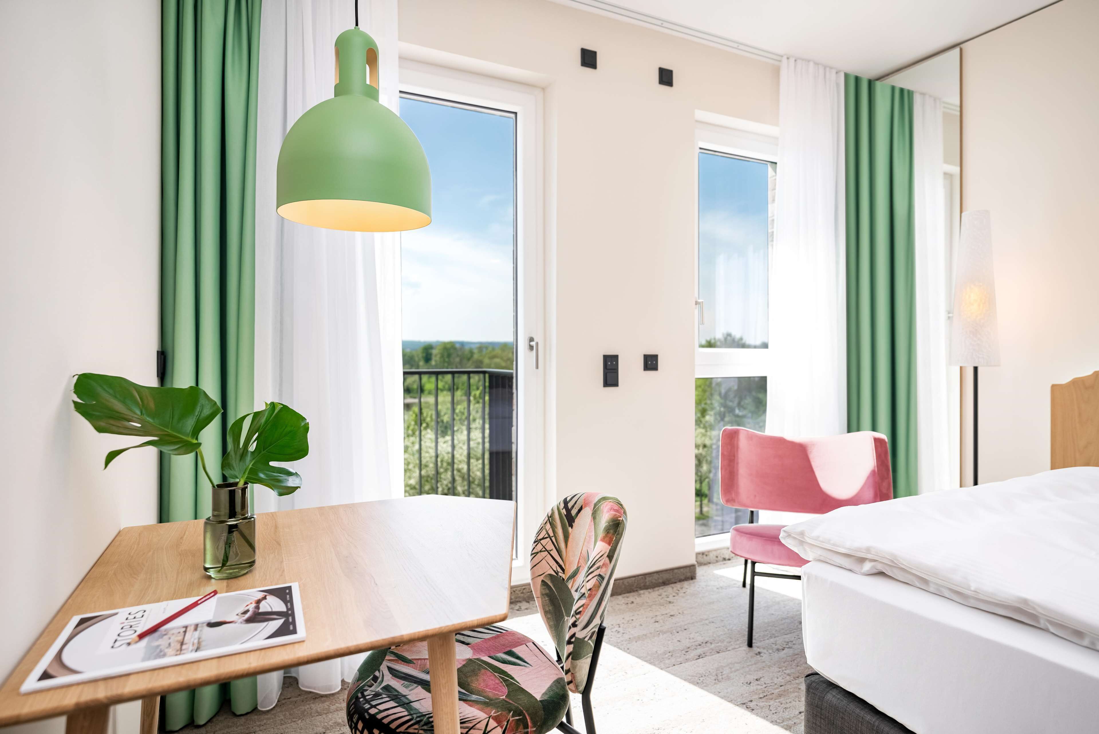 Ein Hotelzimmer mit einem Holztisch, einer grünen Pendelleuchte, einem Stuhl, einem Bett und Blick ins Freie.