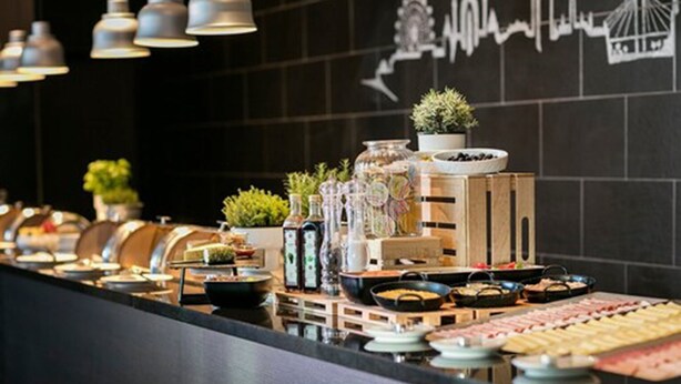 A buffet setup with various food items and condiments, a potted plant, and a wall with a cityscape mural. - ARCOTEL HafenCity Dresden (Dresden)