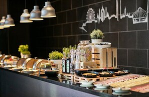 Een buffetopstelling met diverse gerechten en toevoegingen, een potplant en een muur met een stadsgezichtsmural.