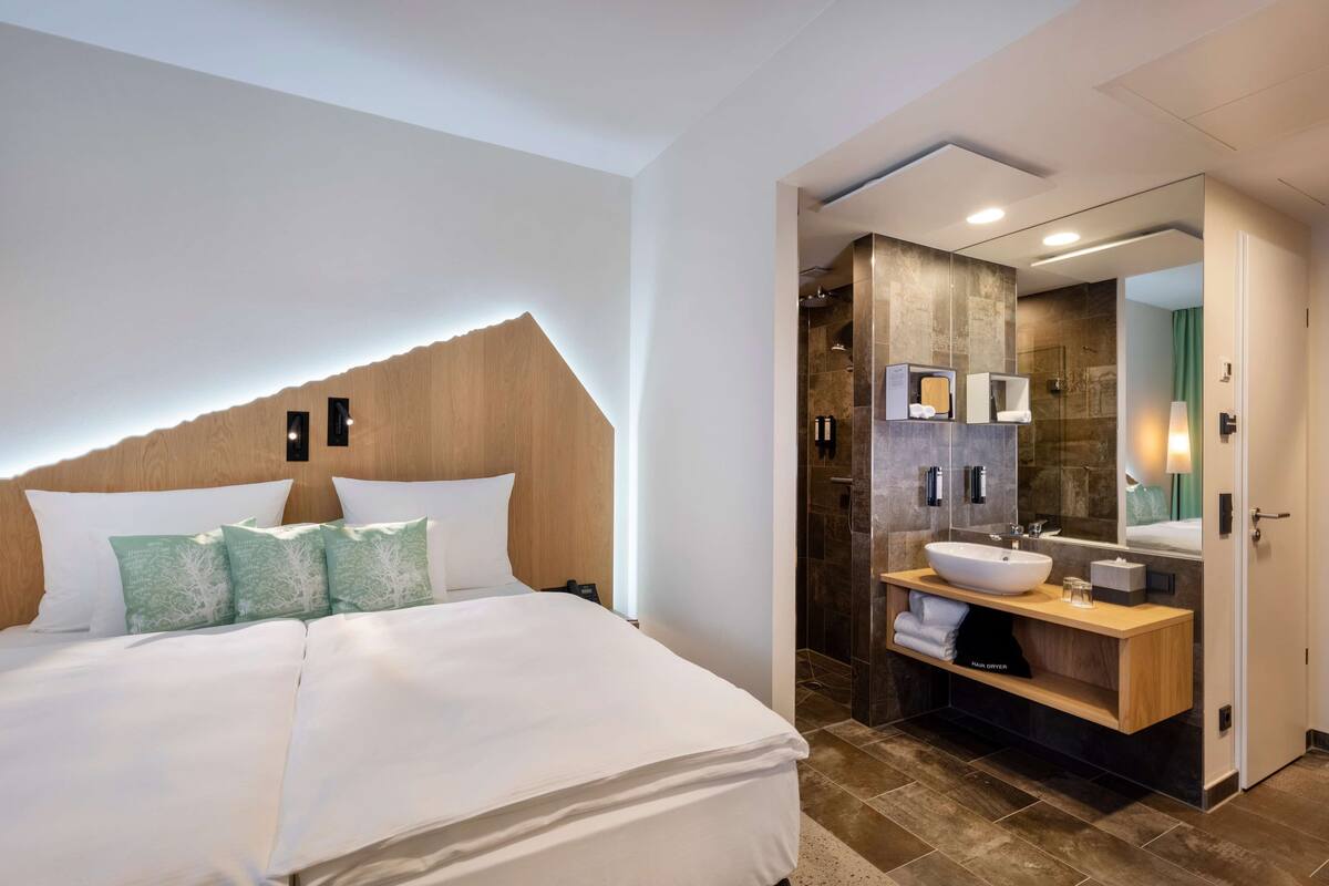 Une chambre d’hôtel moderne avec un grand lit, une tête de lit en bois, une salle de bain dotée d’un grand miroir, d’un lavabo et d’une douche.