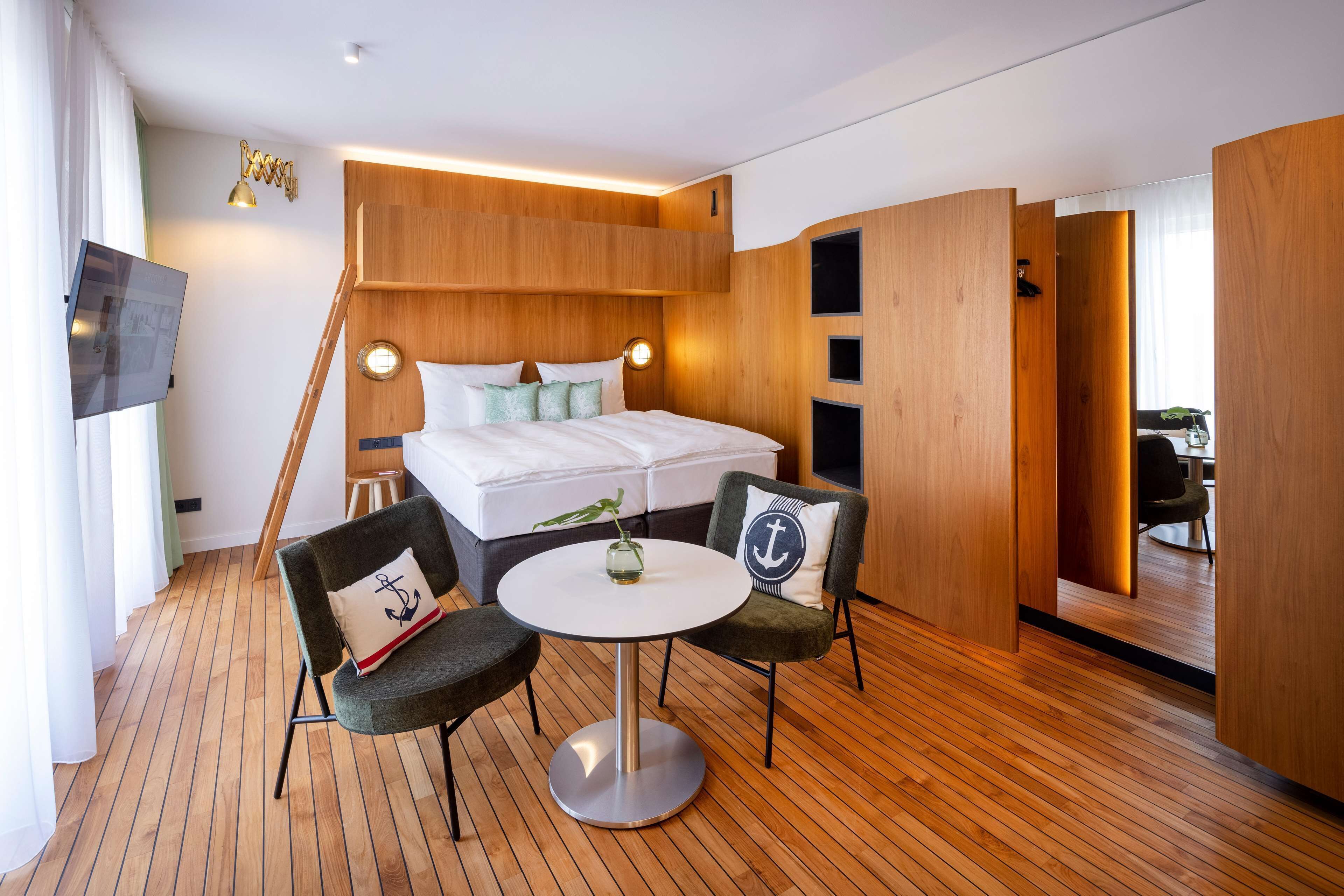 Una habitación de hotel moderna con piso de madera, una cama con sábanas blancas, dos sillones tapizados, una mesa redonda y un televisor montado en la pared.