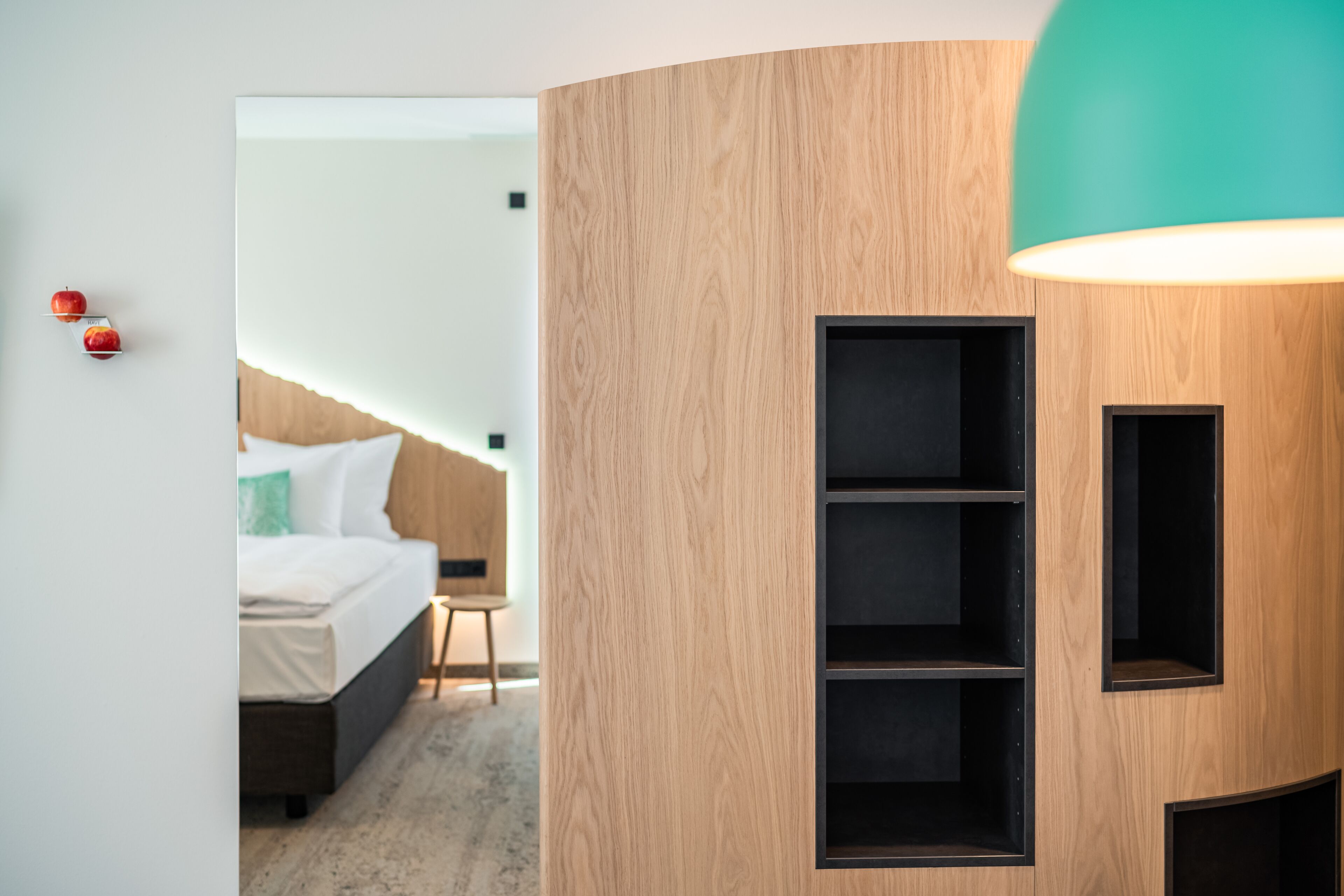 A modern hotel room with a wooden accent wall, a bed with white bedding, and a teal pendant light.
