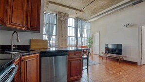 A kitchen with wooden cabinets, a stainless steel dishwasher, a large island, and a TV in a living area with wooden flooring and large windows. - Sosuite | 2BR Apt w W/D, Gym, On-Site Restaurant (Philadelphia)