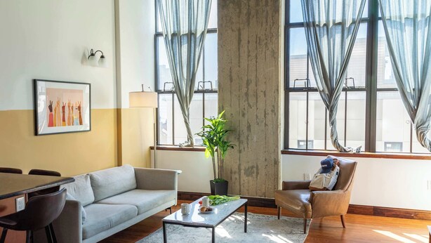 A living room with a sofa, a coffee table, a chair, a plant, and large windows with curtains. - Sosuite | 2BR Apt w W/D, Gym, On-Site Restaurant (Philadelphia)