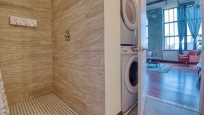A bathroom with a shower, a washing machine, and a view of a living area with a chair and a table. - Sosuite | 2BR Apt w W/D, Gym, On-Site Restaurant (Philadelphia)