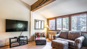 Una sala de estar con un televisor grande, chimenea, ventana con vista a árboles y un rincón de descanso acogedor.