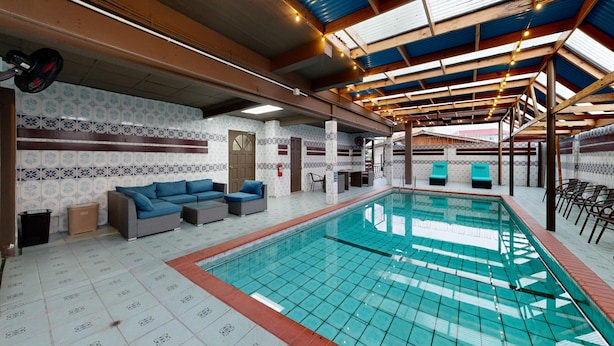 An indoor swimming pool with tiled flooring, blue cushioned seating, and a ceiling with wooden beams and string lights.