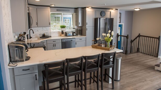 A modern kitchen with a central island, dark wooden chairs, and a stainless steel trash can.