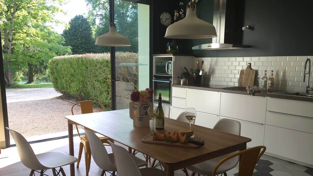 Una cocina moderna con armarios blancos, una mesa de comedor de madera y sillas. Se ve un ventanal con vistas a zonas verdes.