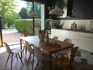 Une cuisine moderne avec des meubles blancs, une table à manger en bois et des chaises. Une fenêtre offre une vue sur la verdure à l’extérieur.