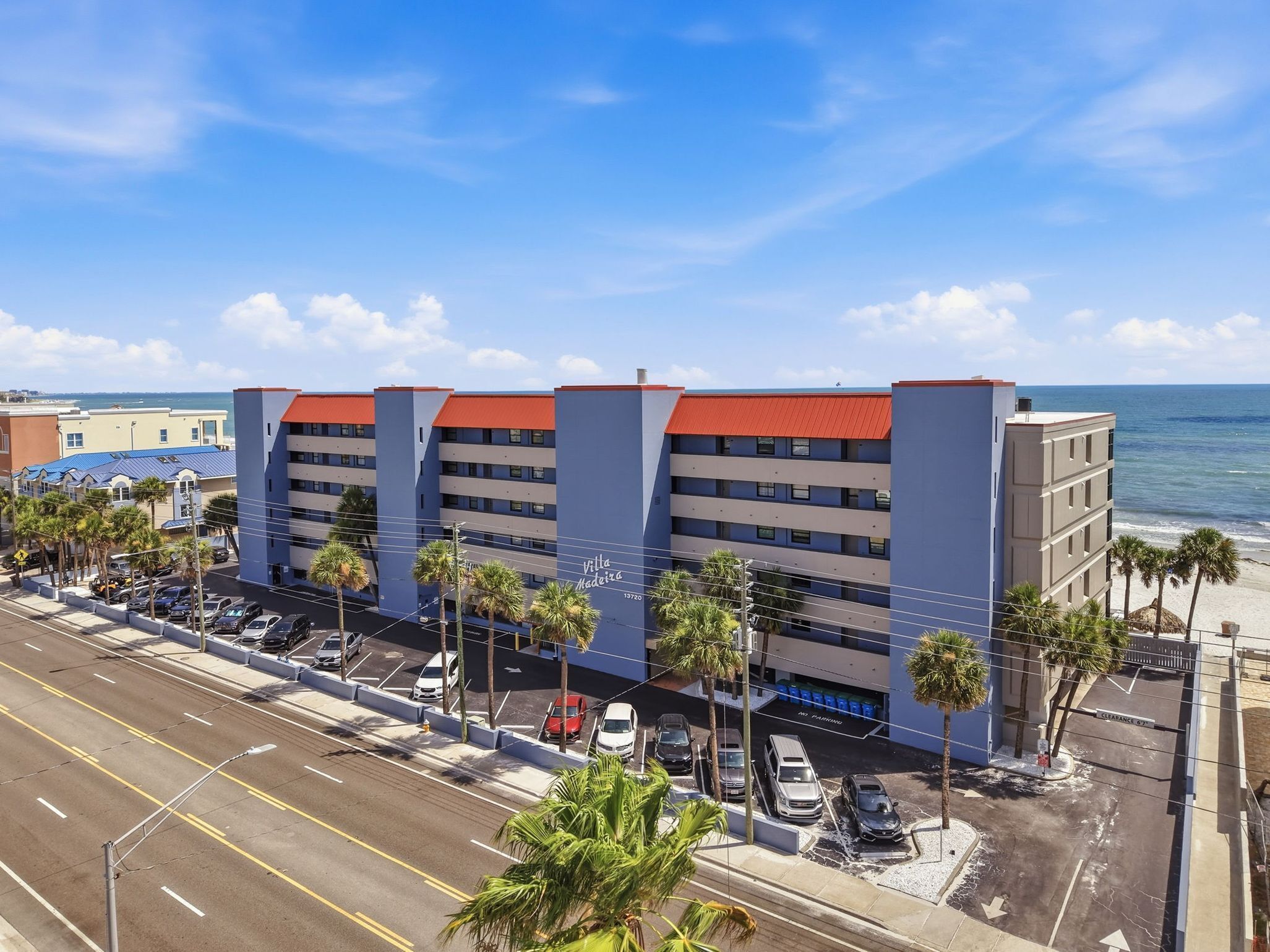 A multi-story hotel with a red roof, located near a beach with palm trees and parked cars.