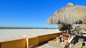 A beachside patio with a thatched umbrella, wooden lounge chairs, and a view of the beach.