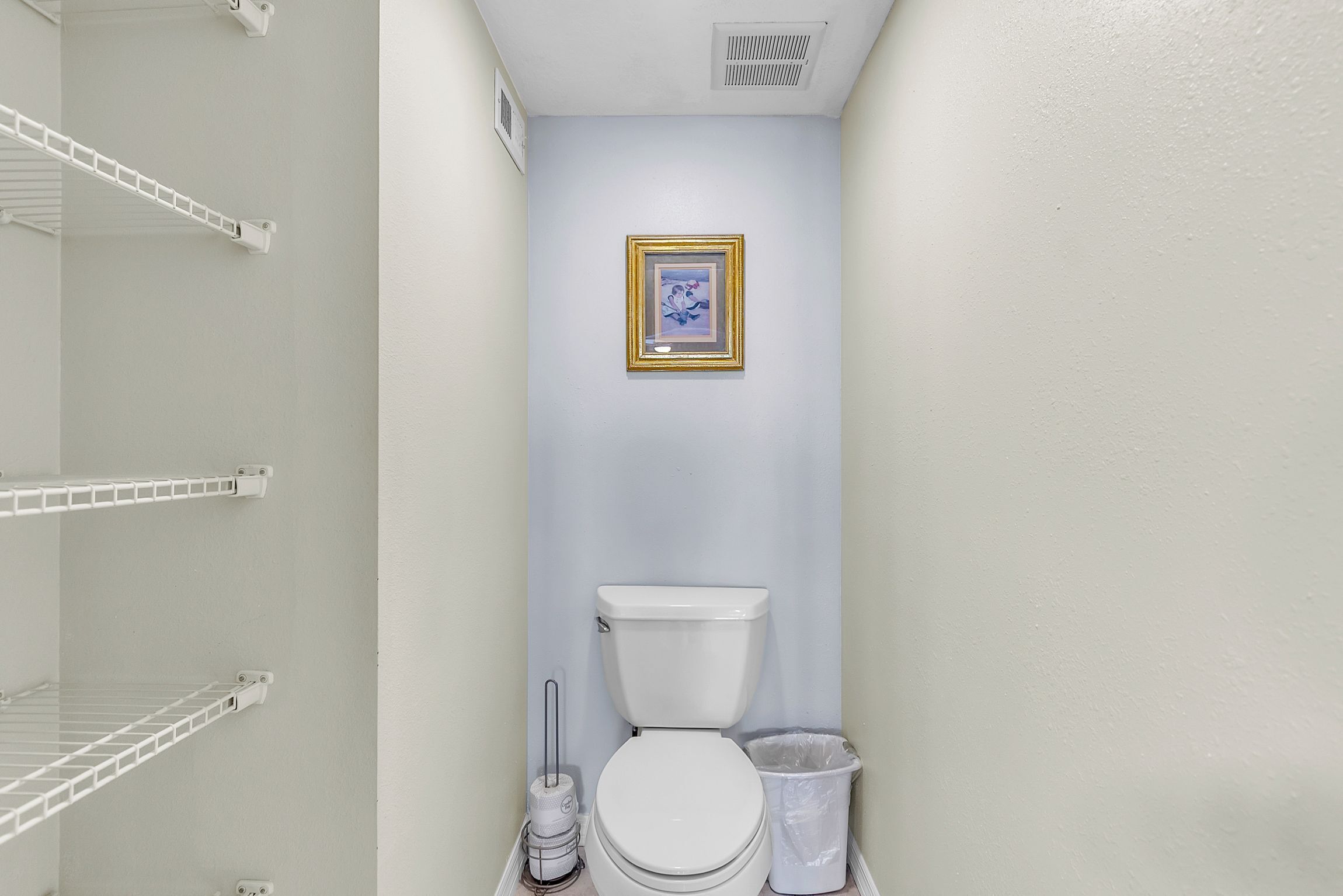 Un bagno con il water, un cestino, un quadro incorniciato a parete e scaffali bianchi.