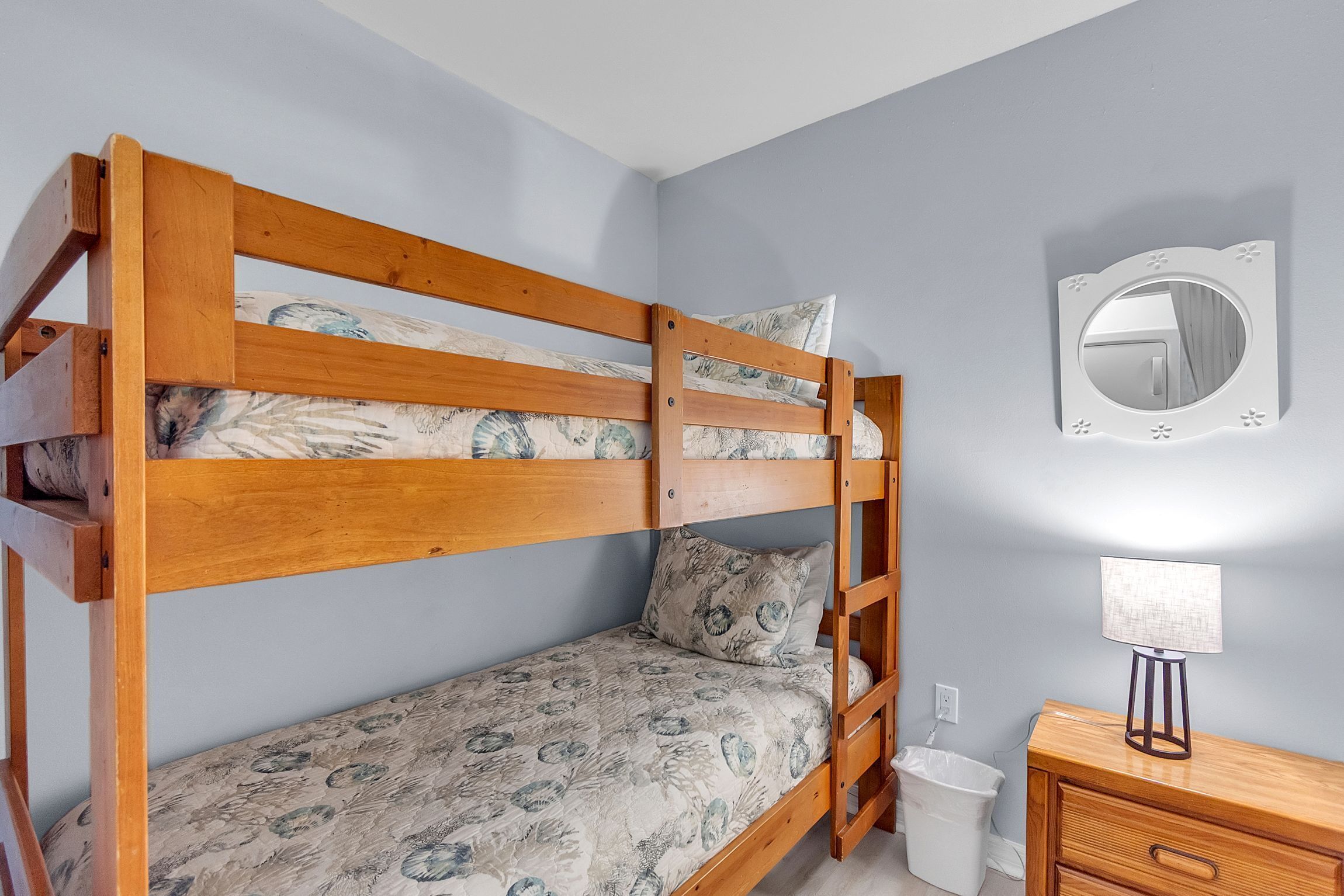 Una cama individual con edredón estampado, una mesita de noche con lámpara y un espejo colgado en la pared.