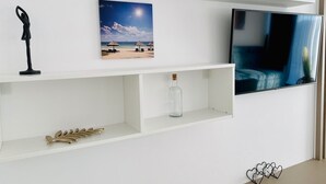 Una pared blanca con un televisor, un cuadro de una playa, una botella y objetos decorativos.