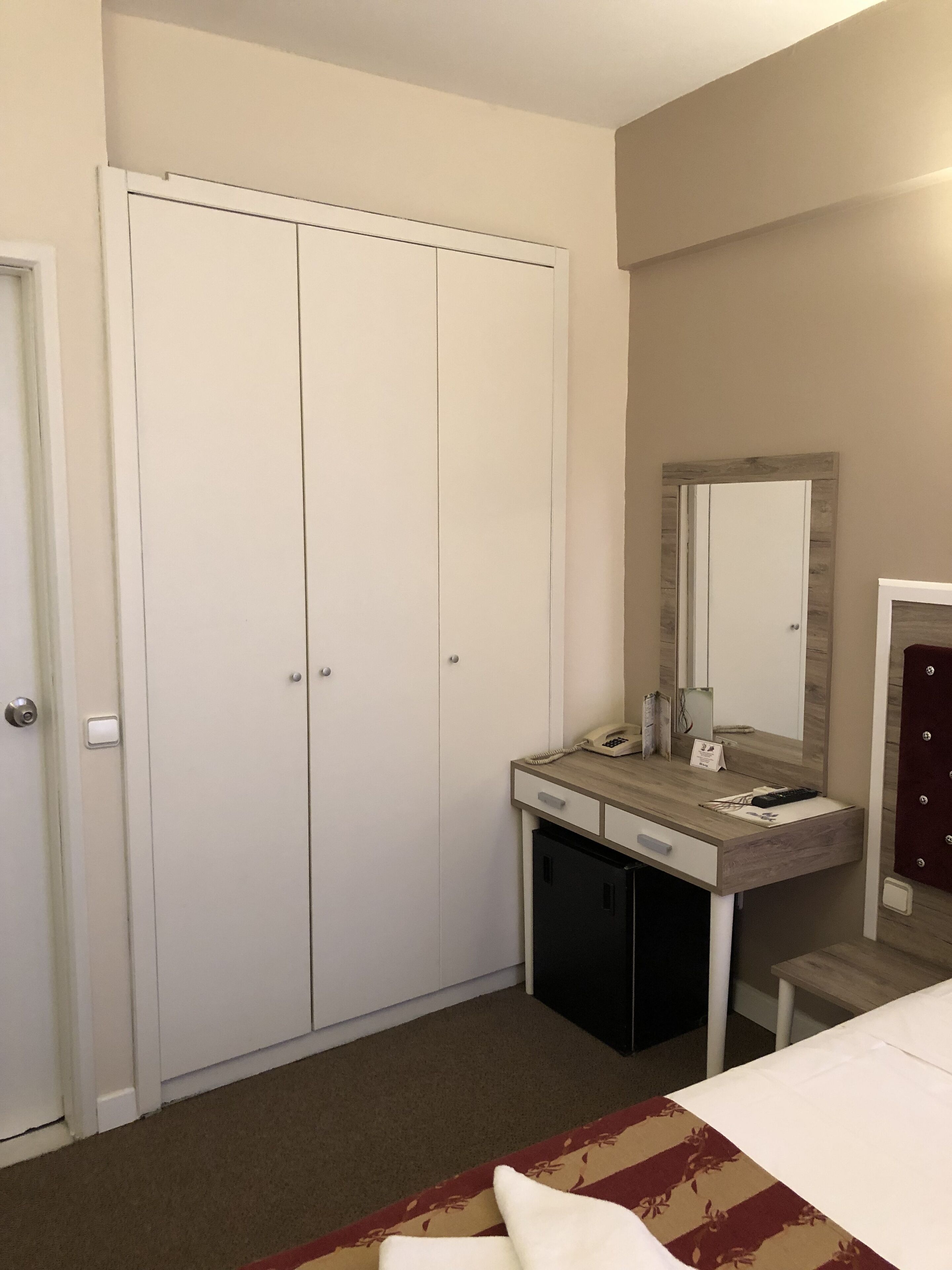A hotel room with a wardrobe, a vanity with a mirror, a desk, and a bed.