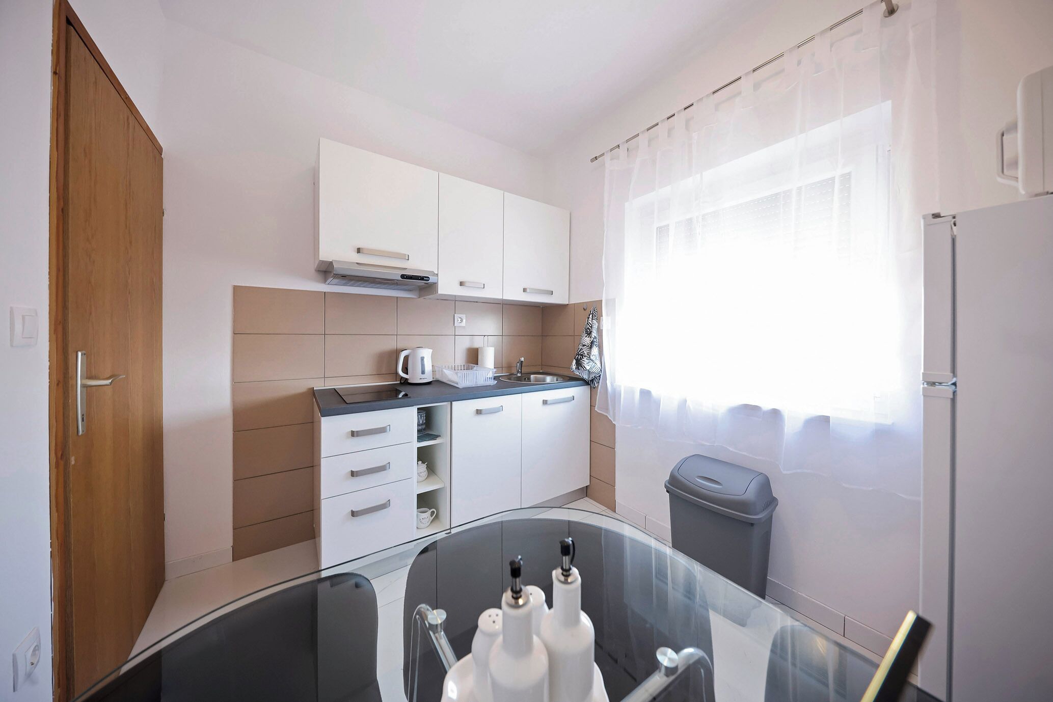 Una cocina moderna con armarios blancos, una mesa de vidrio y una ventana con cortinas translúcidas.
