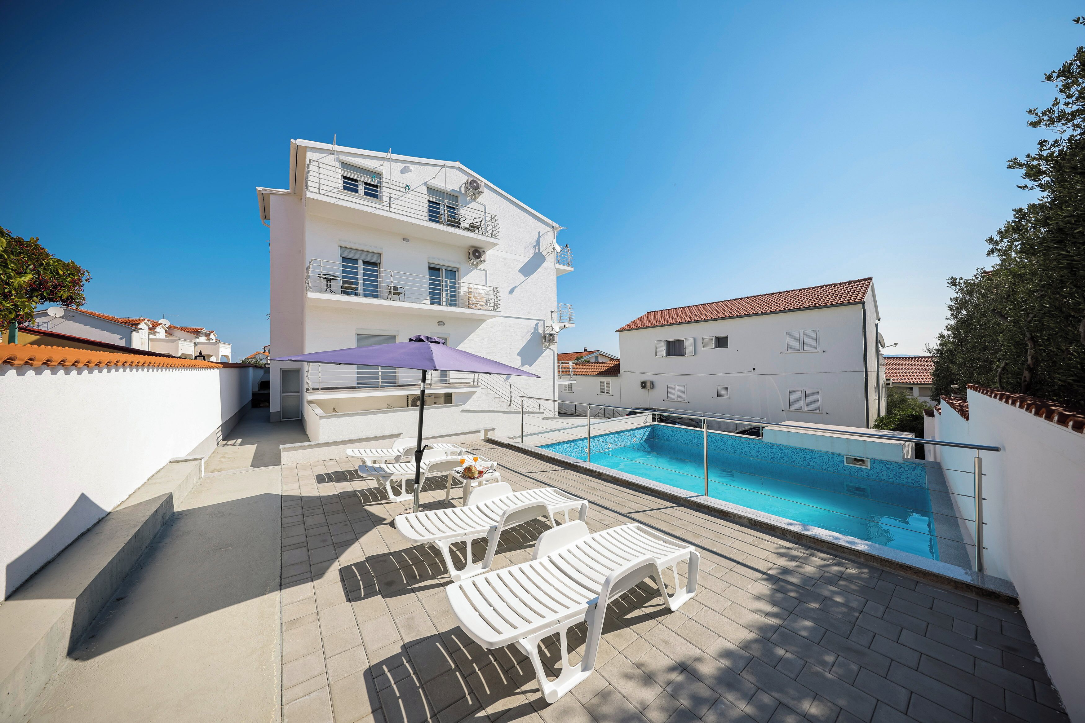 A rooftop pool area with lounge chairs and a table, surrounded by white buildings with balconies.