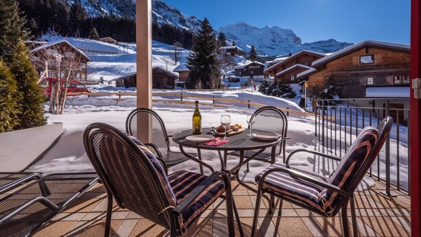 A covered outdoor seating area with a table and chairs, overlooking a snowy mountain landscape with buildings. - Chalet Brunner 2 (Lauterbrunnen)