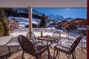 A covered outdoor seating area with a table and chairs, overlooking a snowy mountain landscape with buildings. - Chalet Brunner 2 (Lauterbrunnen)