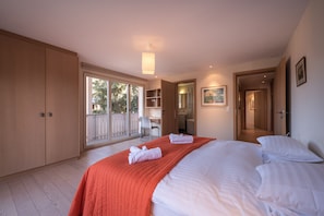 Quarto com uma cama grande, uma escrivaninha e uma cadeira. Há uma porta de vidro deslizante que dá para uma área externa.