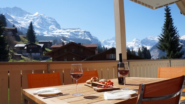 A wooden table set outdoors with a wine glass and bottle, a plate with bread and tomatoes, and a view of snow-capped mountains in the background. - Chalet Brunner 4 (Lauterbrunnen)