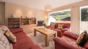 A modern living room with a red sofa set, a wooden coffee table, and a television. There is a bookshelf with books and decorative items, and a view of a house and hills through a large window. - Chalet Brunner 4 (Lauterbrunnen)