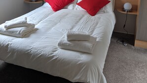 A neatly made bed with white linens and red pillows, a bedside table with a lamp, and a wooden shelf above the bed. - Caravan Nearby Hastings, Coghurst Hall Park (Hastings)