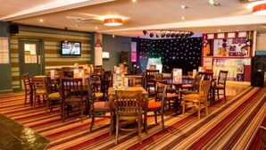A restaurant with striped carpeting, wooden tables and chairs, a stage with lights, and a TV on the wall. - Caravan Nearby Hastings, Coghurst Hall Park (Hastings)