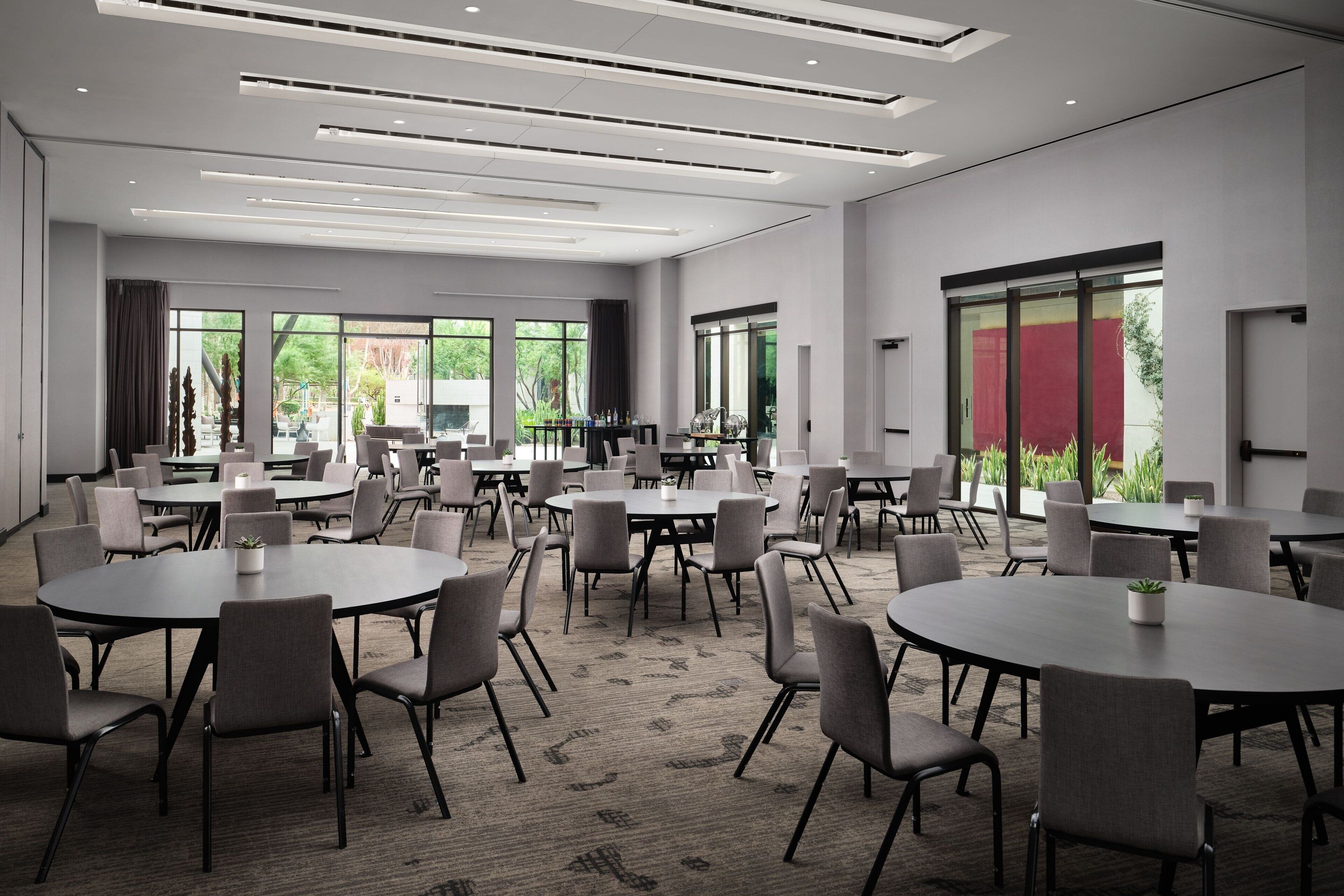 Ein moderner Konferenzraum mit mehreren runden Tischen und Stühlen, großen Fenstern mit Blick auf Grünflächen und einem klaren, minimalistischen Design.