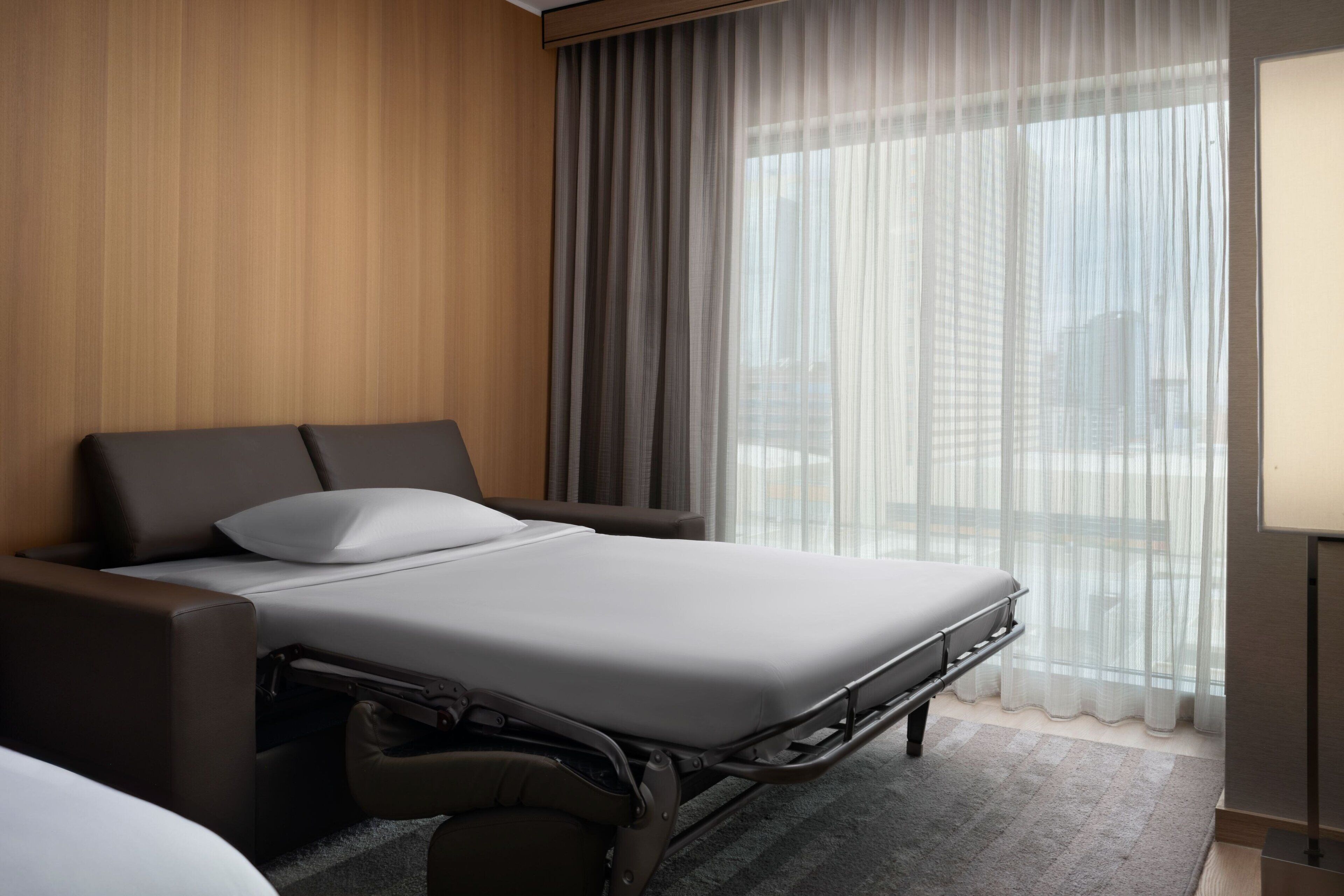 Ett modernt hotellrum med en säng, ett sängbord och ett fönster med tunna, genomskinliga gardiner.