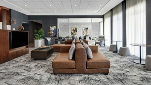 A modern hotel lobby with a large sectional sofa, a flat-screen TV, and a coffee table. There are multiple seating areas and a large window with curtains. - Courtyard by Marriott Hamilton (Robbinsville)