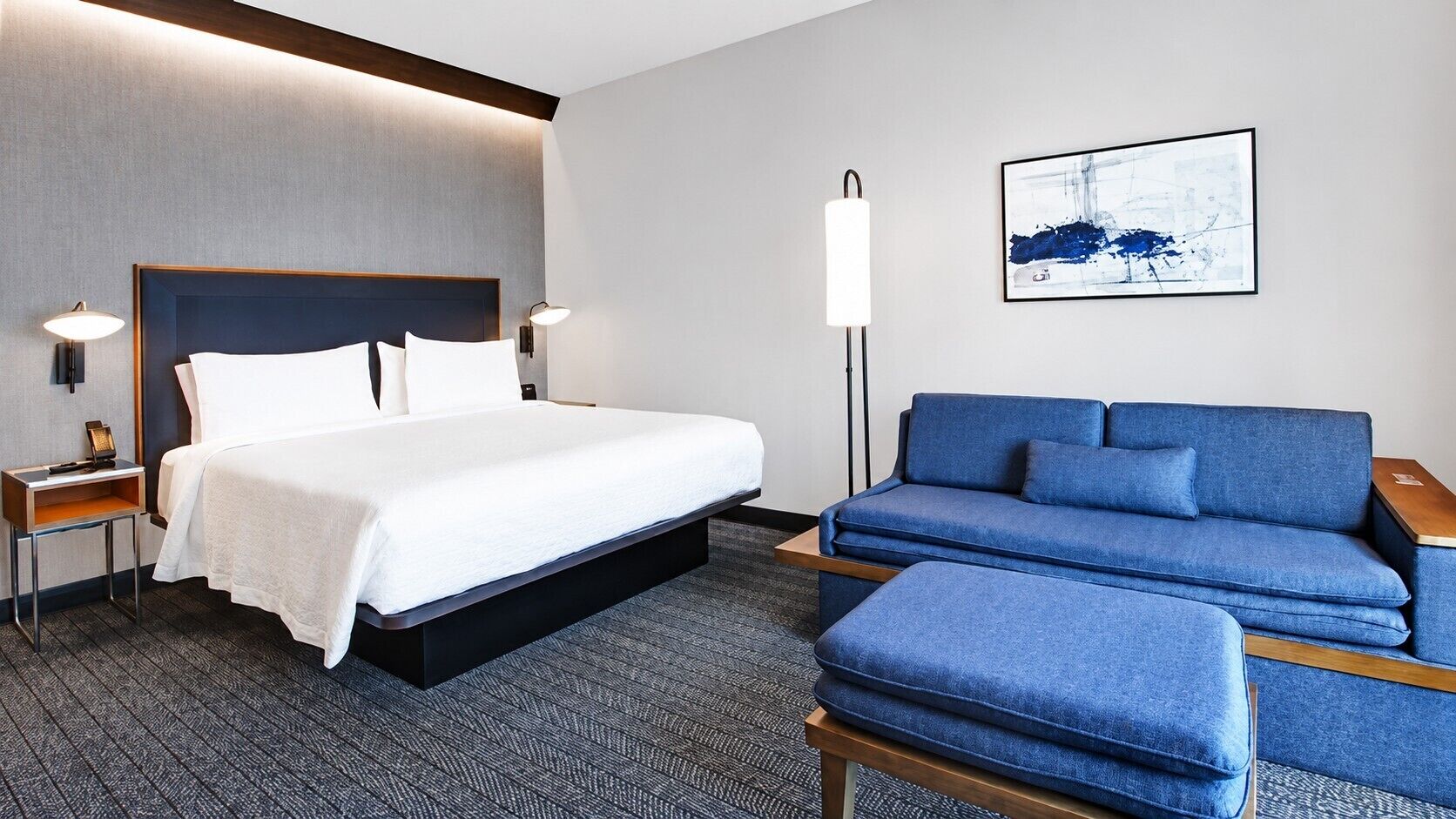 Una habitación de hotel con una cama, un sofá azul, una lámpara de pie y un cuadro en la pared.
