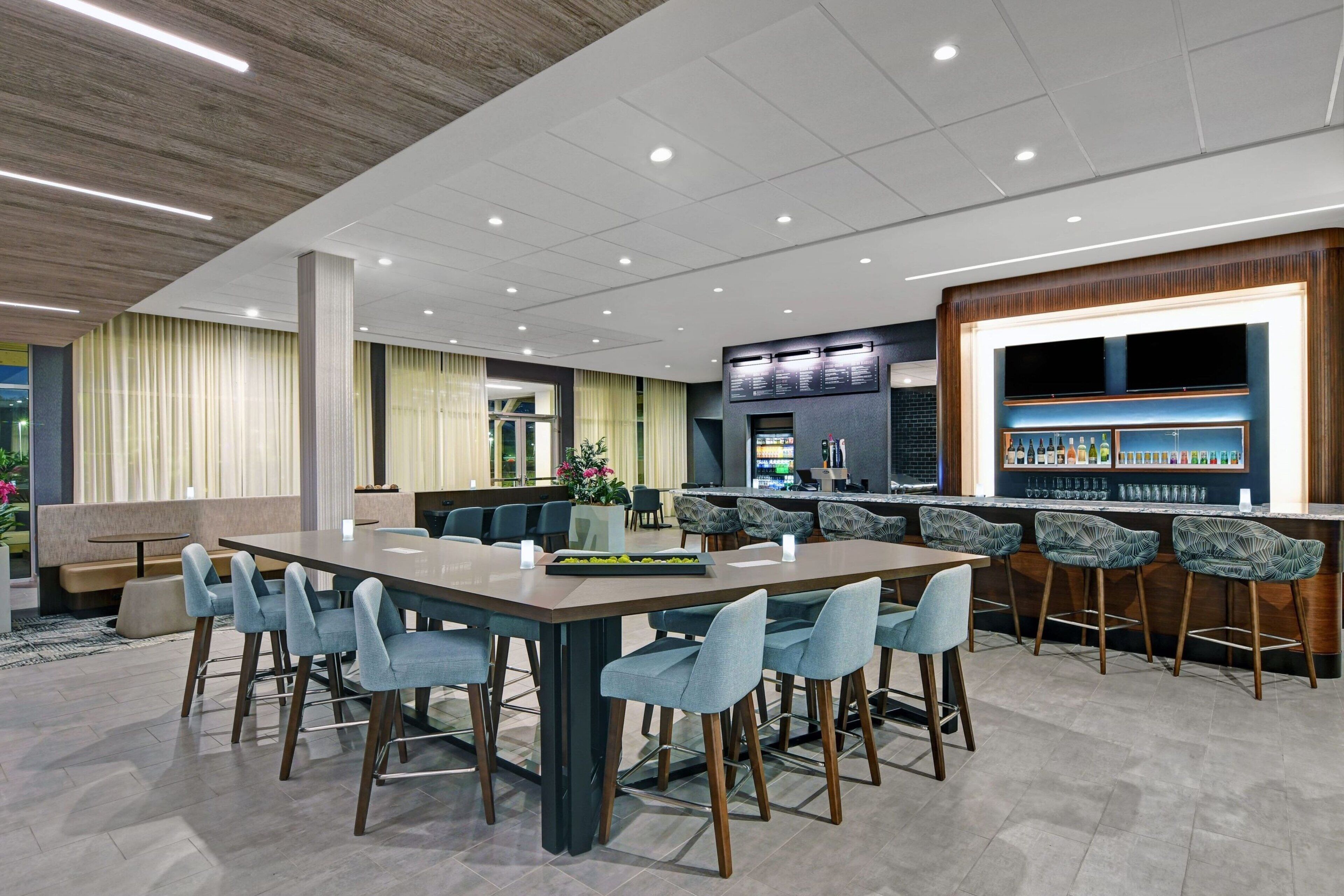 A modern dining area with a long table and multiple chairs, a bar with stools, and a kitchen area in the background.